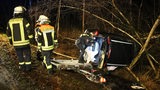 Ein Auto liegt in einem Graben. Eine Polizistin schaut hinein. Daneben stehen Feuerwehrleute.