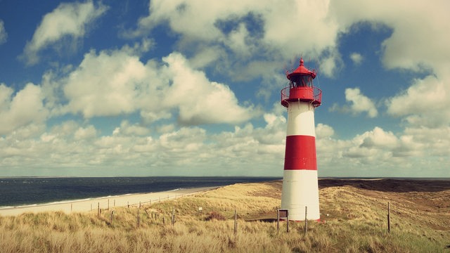 Ein rot-weißer Leuchtturm zwischen Dünen. Dahinter ein blauer Himmel mit ein paar Wolken. 