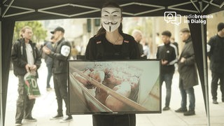 Ein Mann mit Maske hält ein Foto hoch mit vielen Schweinen in einem kleinen Stall.